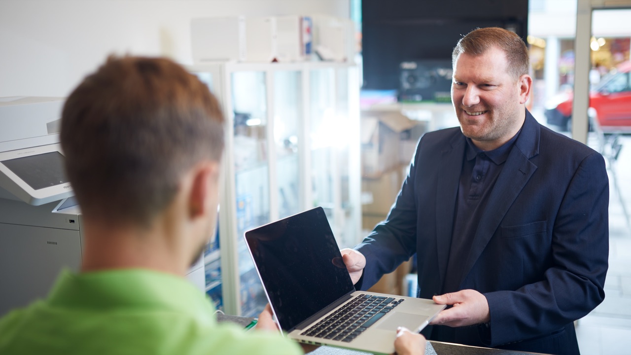 Kunde nimmt lächelnd sein MacBook am Tresen entgegen, Mitarbeiter von hinten